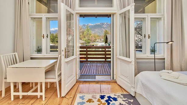 A room with a bed, a desk, and chairs, a window with a view of mountains, and a balcony with a wooden deck.