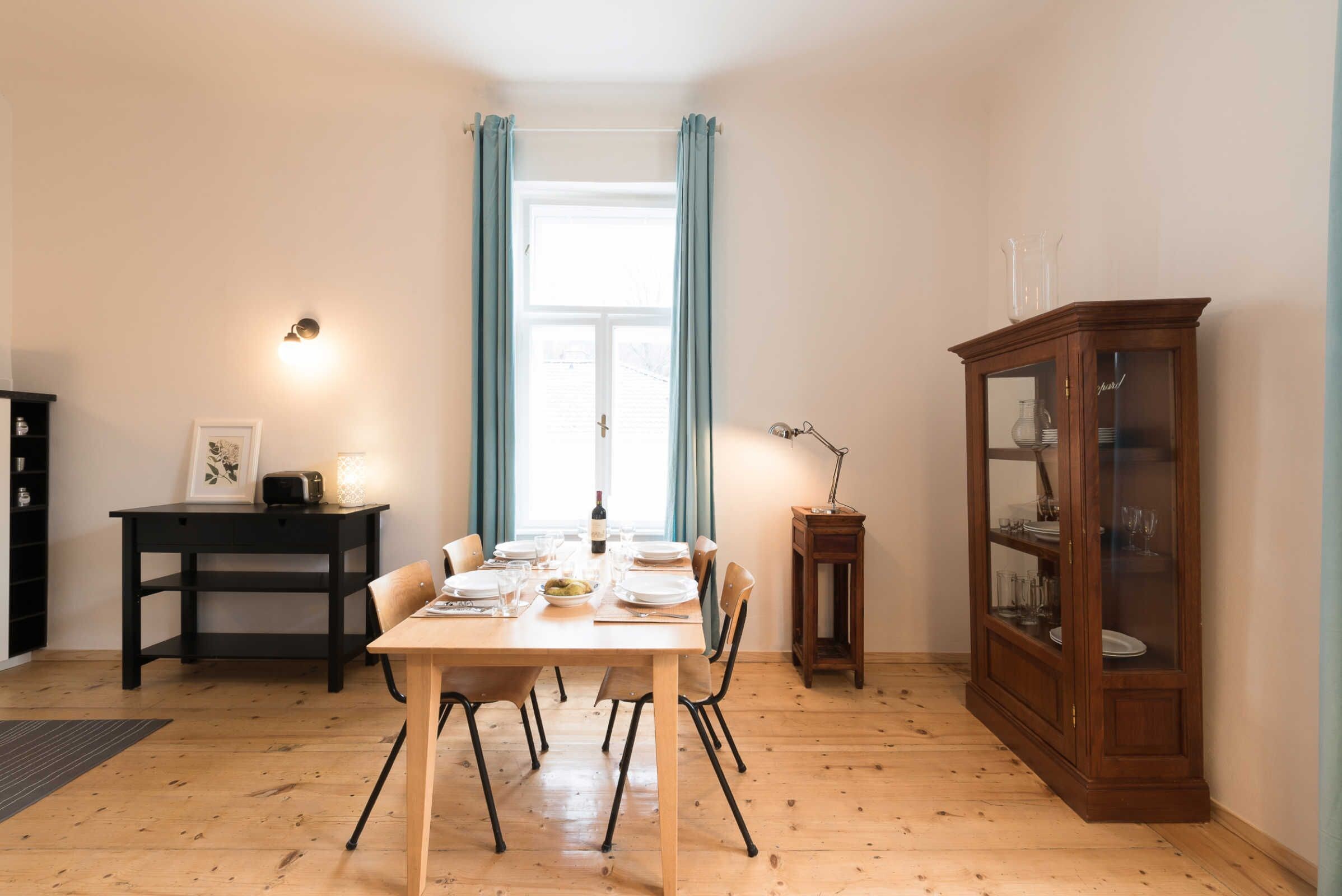 A dining area with a wooden table set for six, a cabinet with glass doors, and a window with curtains.