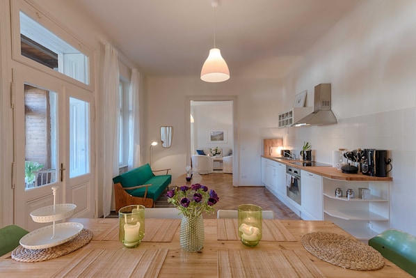 A modern kitchen with a dining area, featuring a wooden table, a hanging pendant light, and a view into the living room.