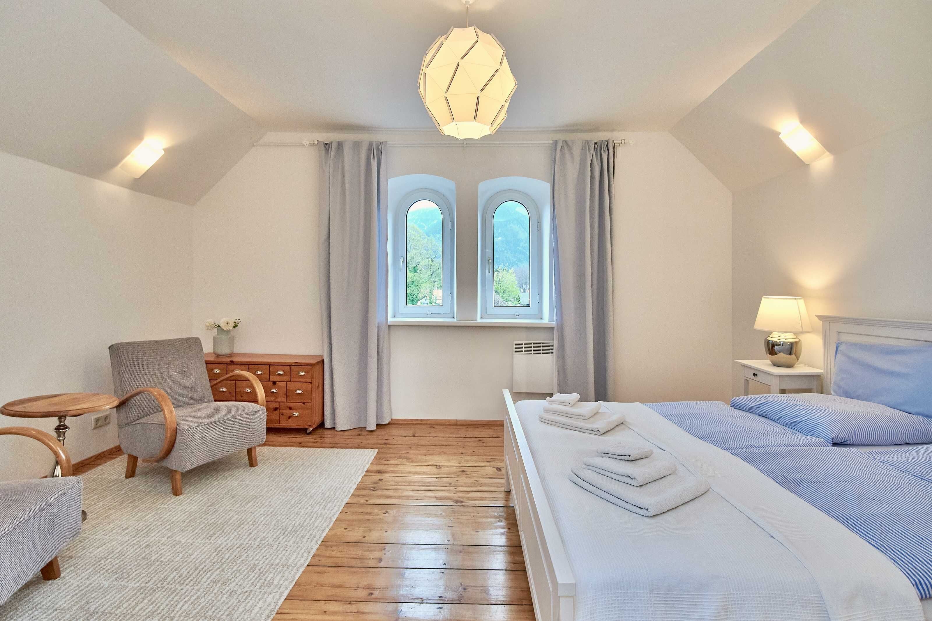 A bedroom with a bed, a chair, a small table, a dresser, and two windows with curtains.