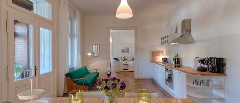A modern kitchen with a dining area, featuring a wooden table, a hanging pendant light, and a view into the living room.