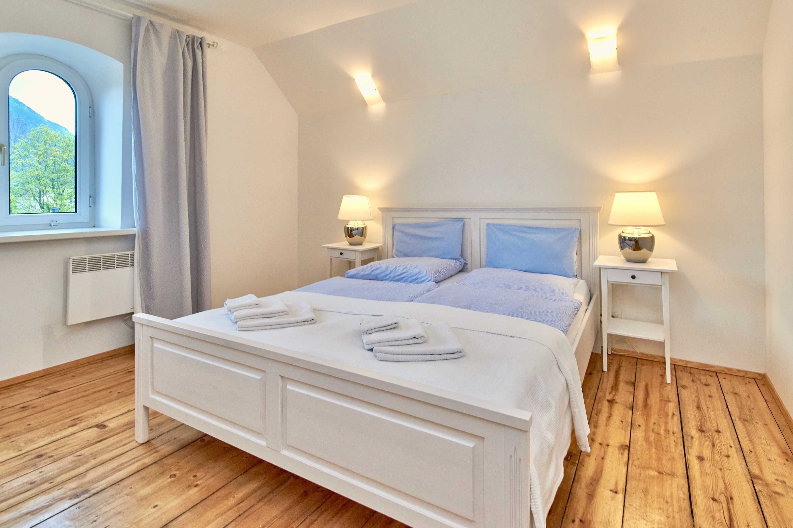 A bedroom with a bed, two bedside tables, a window with curtains, and a view of trees.