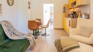 A modern living room with a green sofa, a beige armchair, and a dining area with two chairs. A kitchen with yellow cabinets and a microwave is visible in the background. - Duggis Feriendomizil - Ferienwohnung (Wolgast)