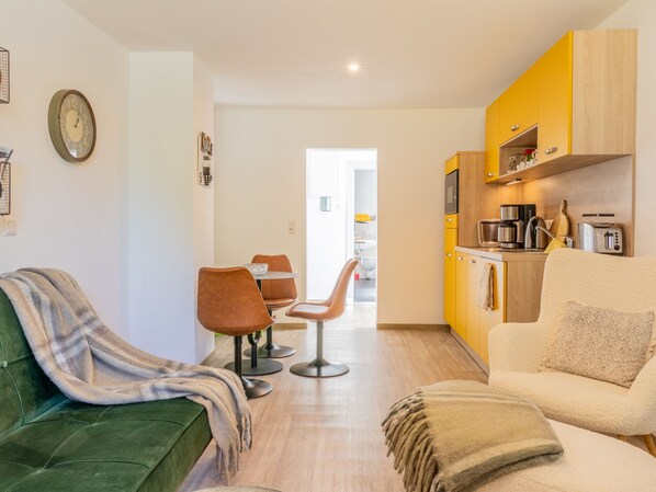 Un salon moderne avec un canapé vert, un fauteuil beige et un coin repas avec deux chaises. On aperçoit, au fond, une cuisine avec des placards jaunes et un four à micro-ondes.