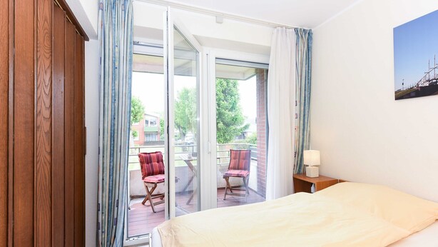 Een hotelkamer met een bed, nachtkastje, stoel en een balkon met gordijnen.