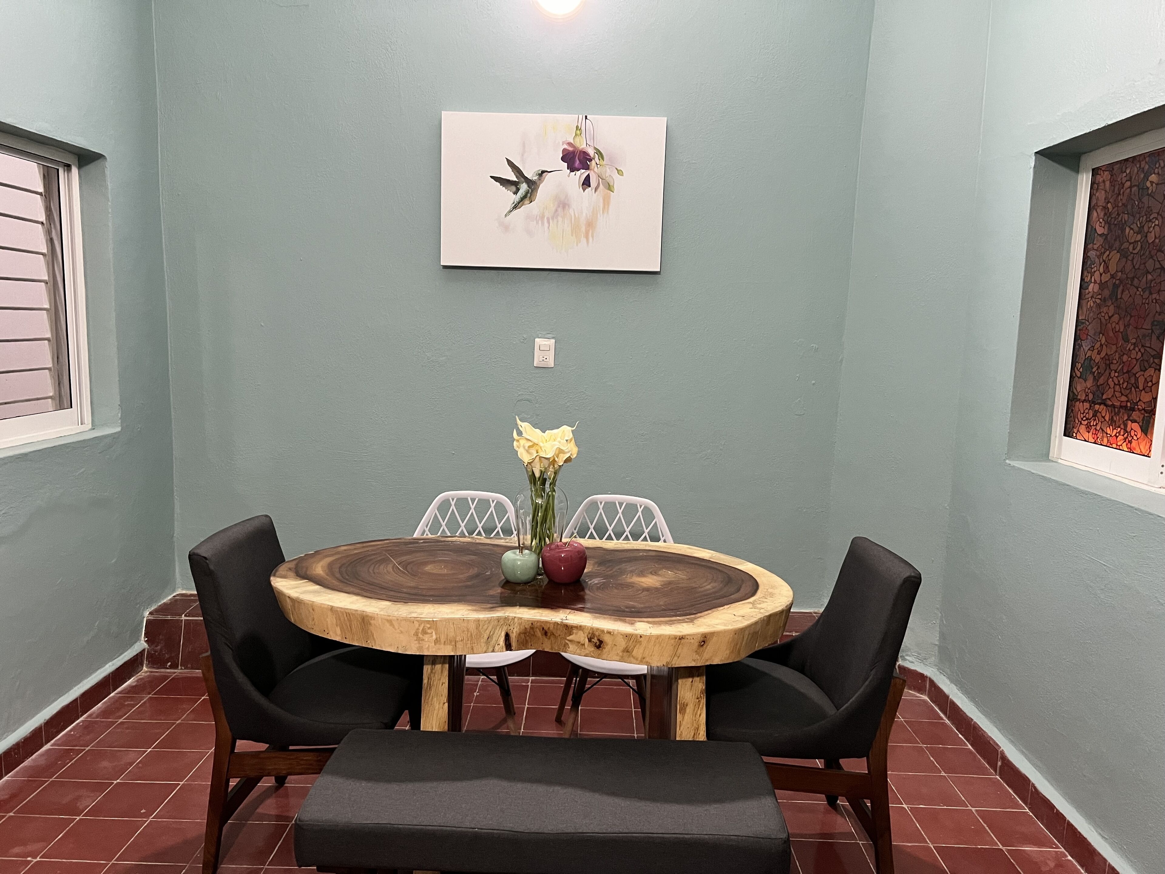 A dining area with a round wooden table, black chairs, and a bench. There is a painting on the wall above the table.
