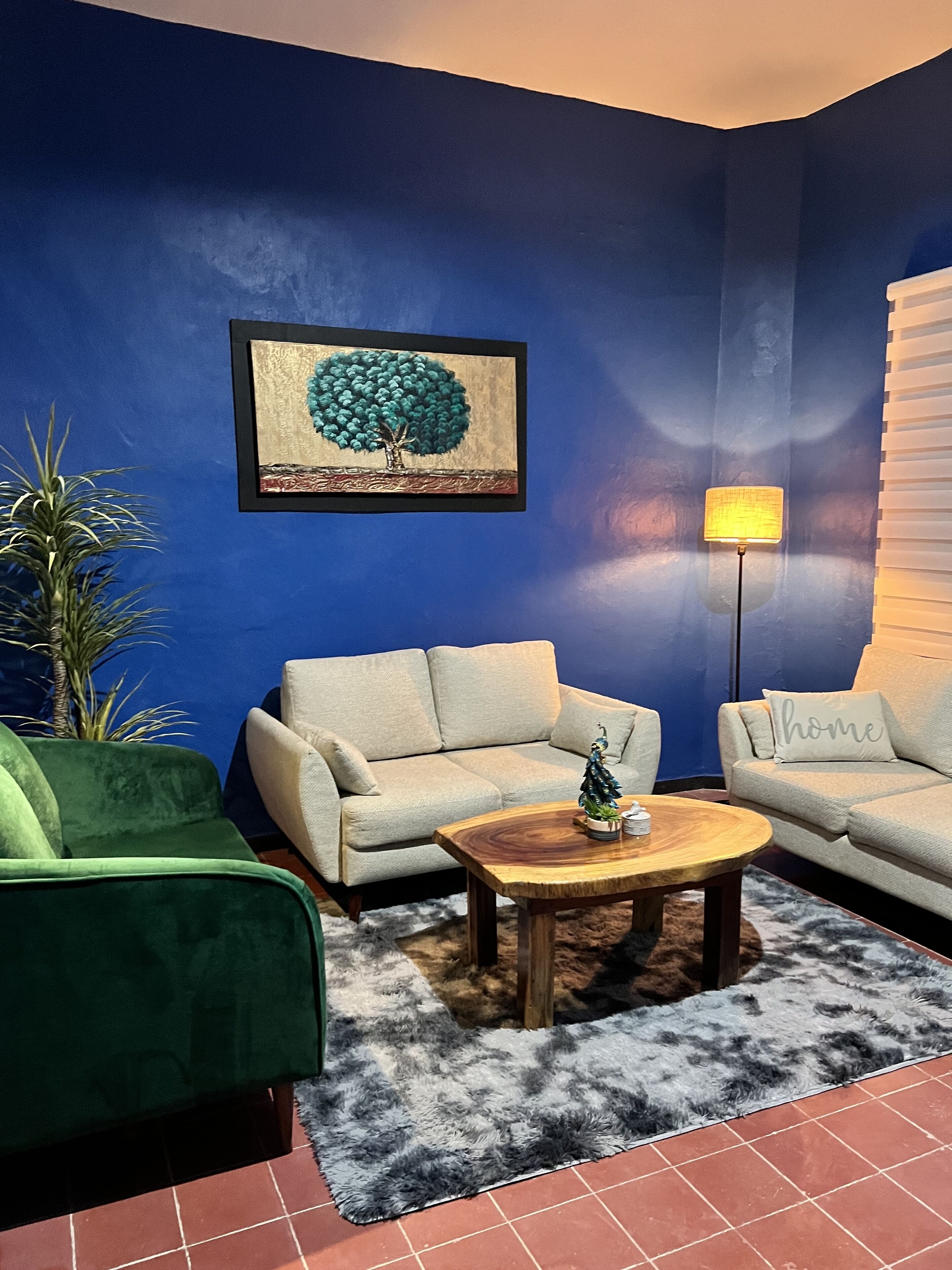 A living room with a blue wall, two light-colored sofas, a green armchair, a wooden coffee table, a small Christmas tree, and a lamp.