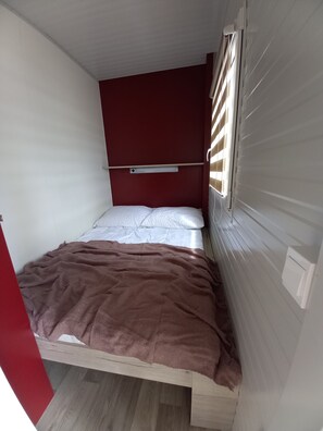 A small, single-bed room with a wooden frame, a red headboard, and a window with blinds. - Sun and Sand Resort Mobile Home, Privlaka (Privlaka)