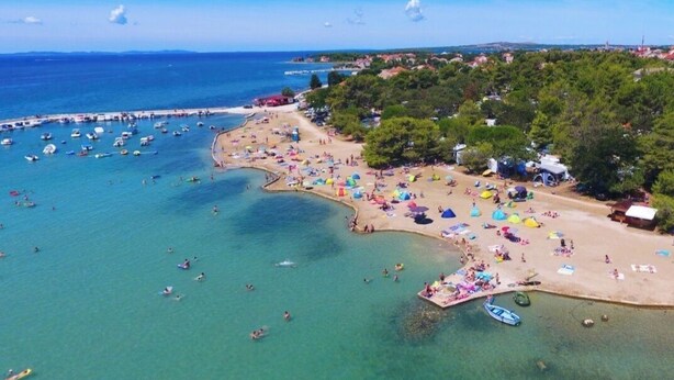 Vista aérea de una playa con aguas cristalinas, numerosos barcos y gente disfrutando de la playa y actividades acuáticas.