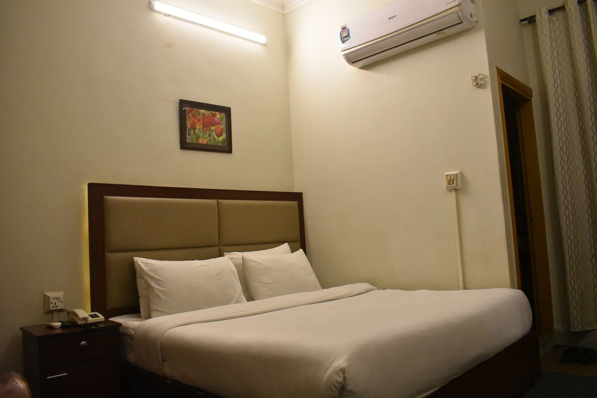 A hotel room with a bed, a nightstand, an air conditioning unit, and a door.