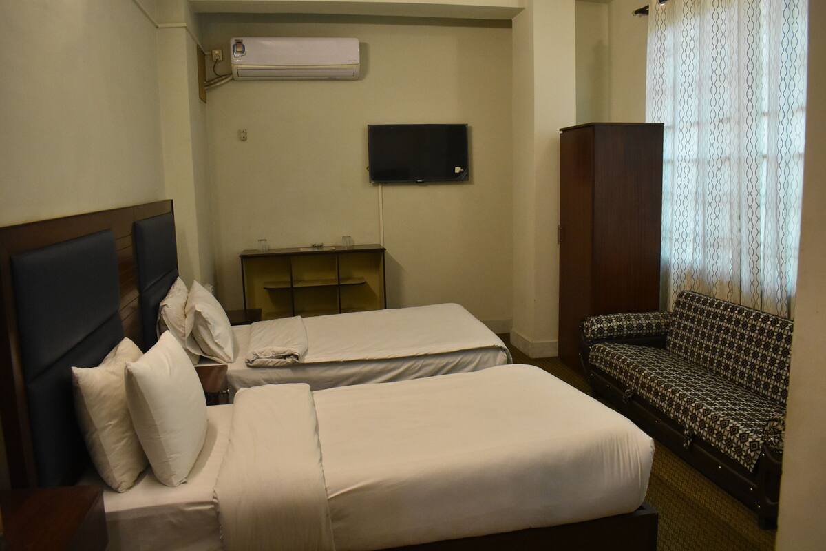A hotel room with two beds, a TV, a sofa, and an air conditioning unit.