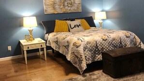 Ein Schlafzimmer mit einem Bett, zwei Nachttischen mit Lampen, einem kleinen Tisch, einem Zierkissen, einem Wandbild und einem Koffer.