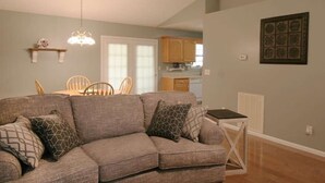 Ein Wohnzimmer mit grauem Sofa, gemusterten Kissen, einem Essbereich mit Tisch und Stühlen sowie einer Küche im Hintergrund.