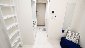 A narrow hallway with white tiled flooring, a white door at the end, and a blue bench with patterned cushions. - Studio G2 (Tokyo)