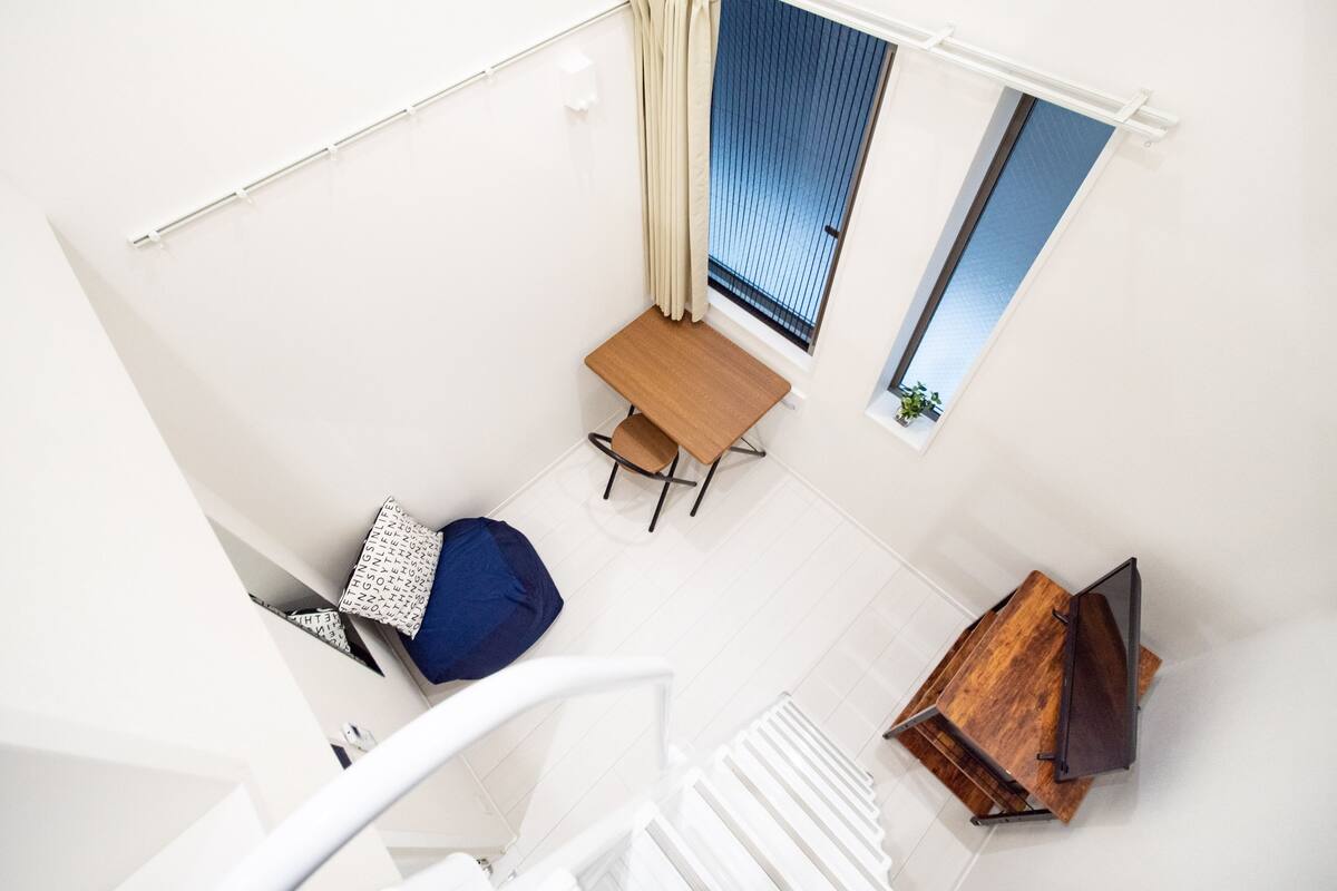 An aerial view of a room with a wooden table and chairs, a blue bean bag, a small window with a view, and a white staircase.