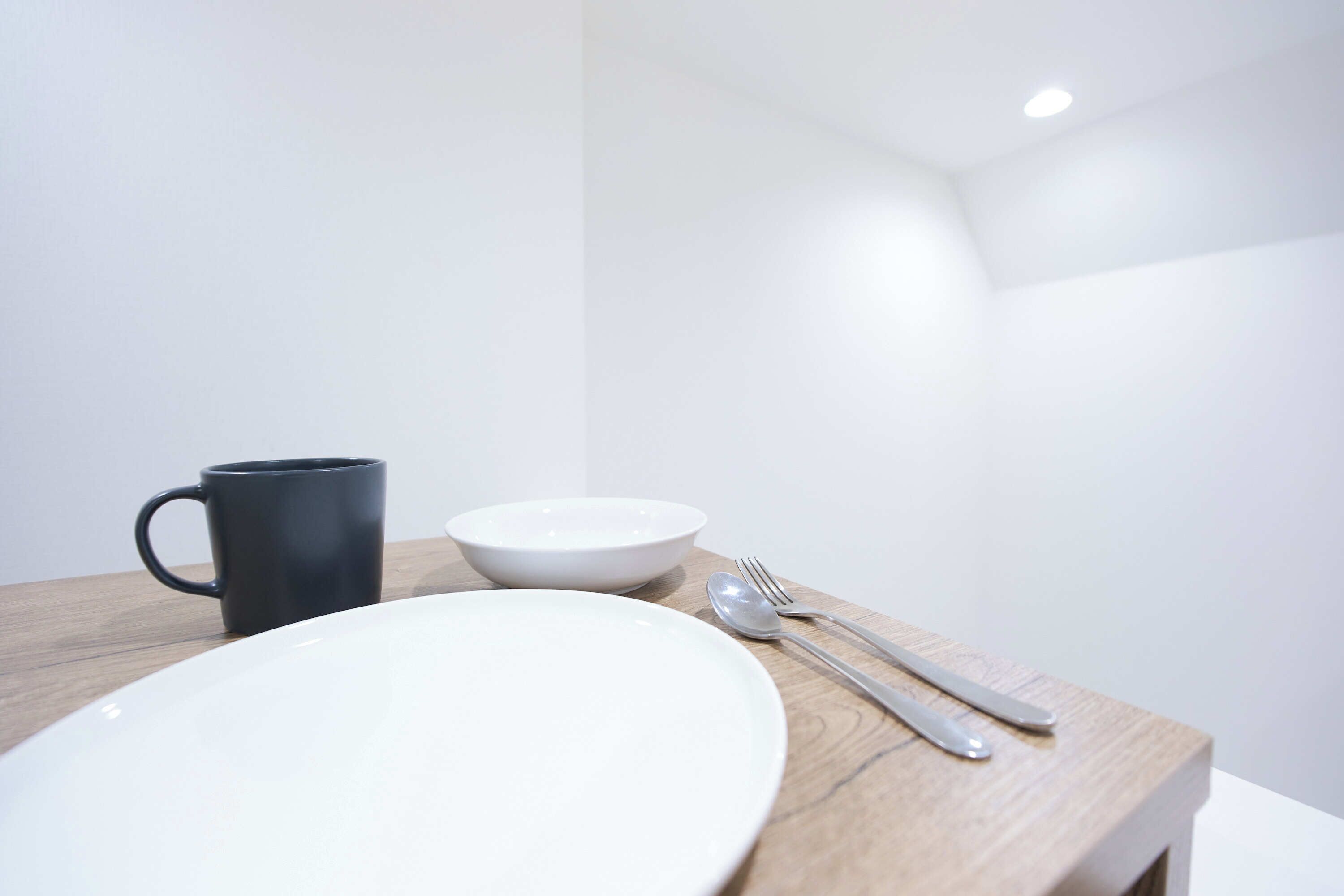 Disposição minimalista para refeições, com um prato branco, uma caneca preta e uma tigela branca sobre uma mesa de madeira.