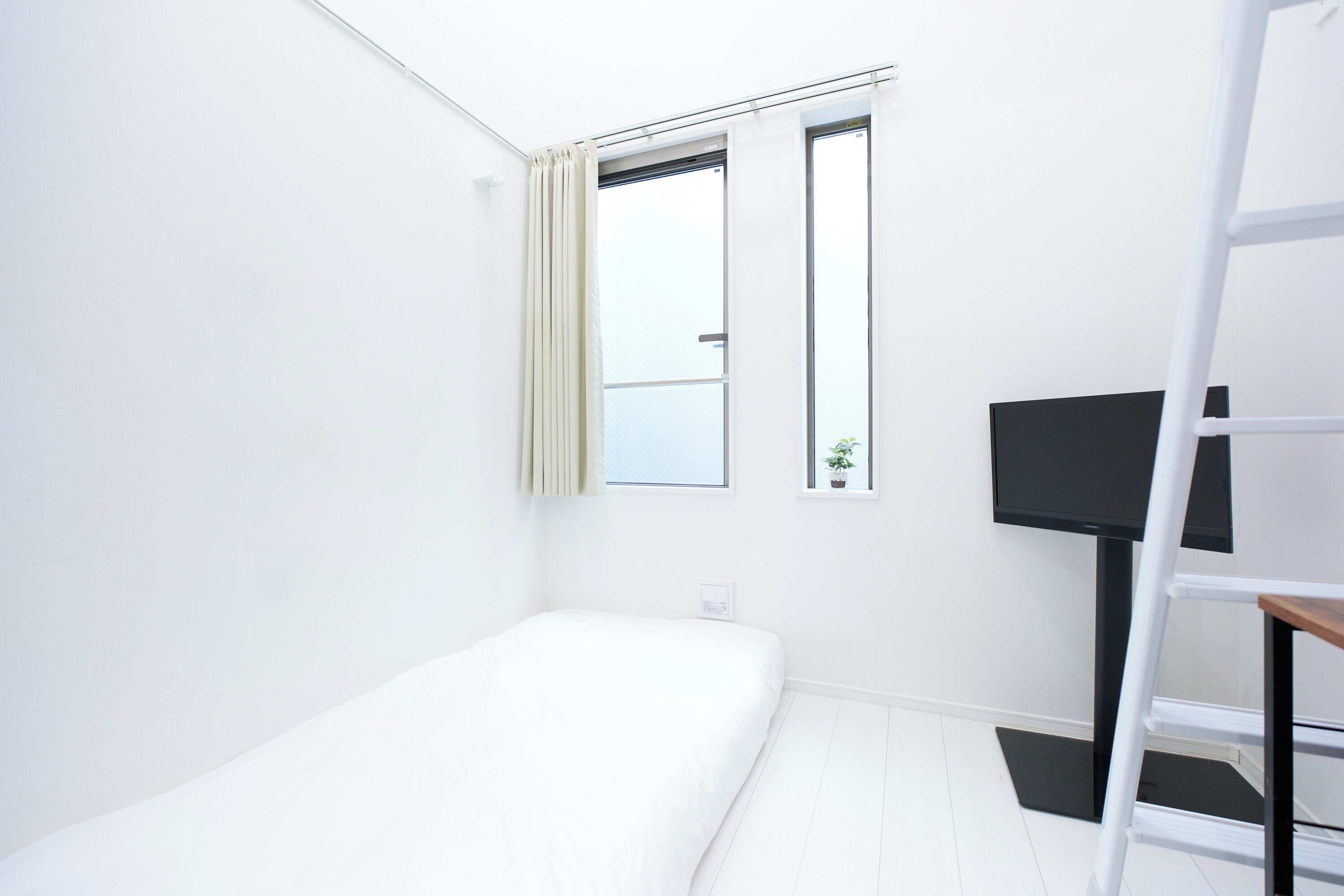 Quarto pequeno e luminoso com uma cama de solteiro, janela com cortinas, uma mesa pequena com uma planta e uma TV sobre um suporte.