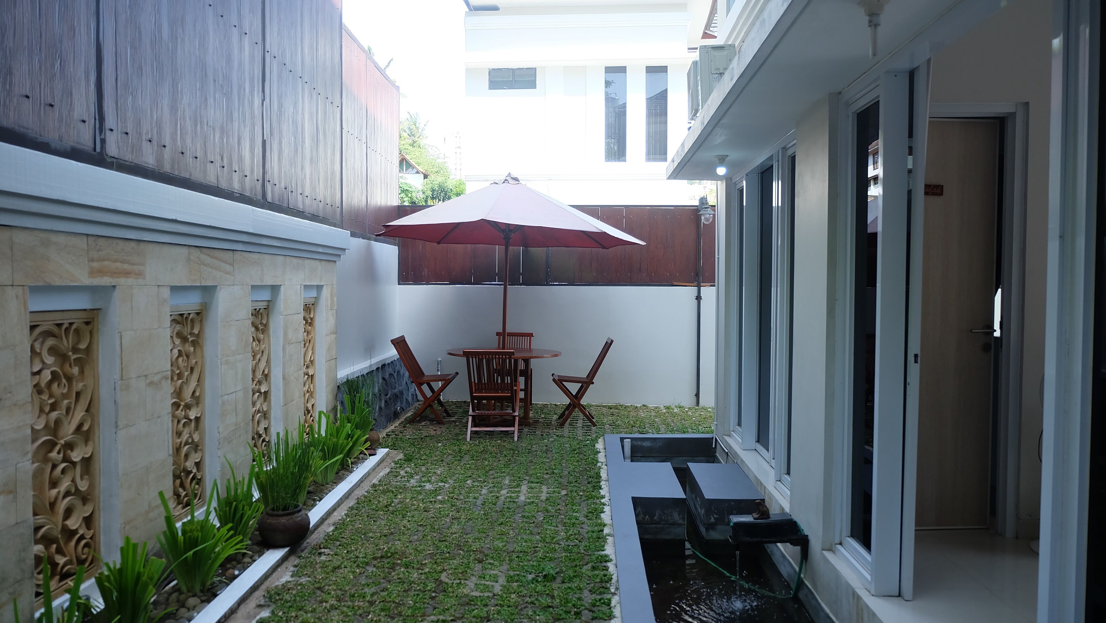 A small, enclosed patio with a table and chairs under a red umbrella, surrounded by decorative stonework and plants.
