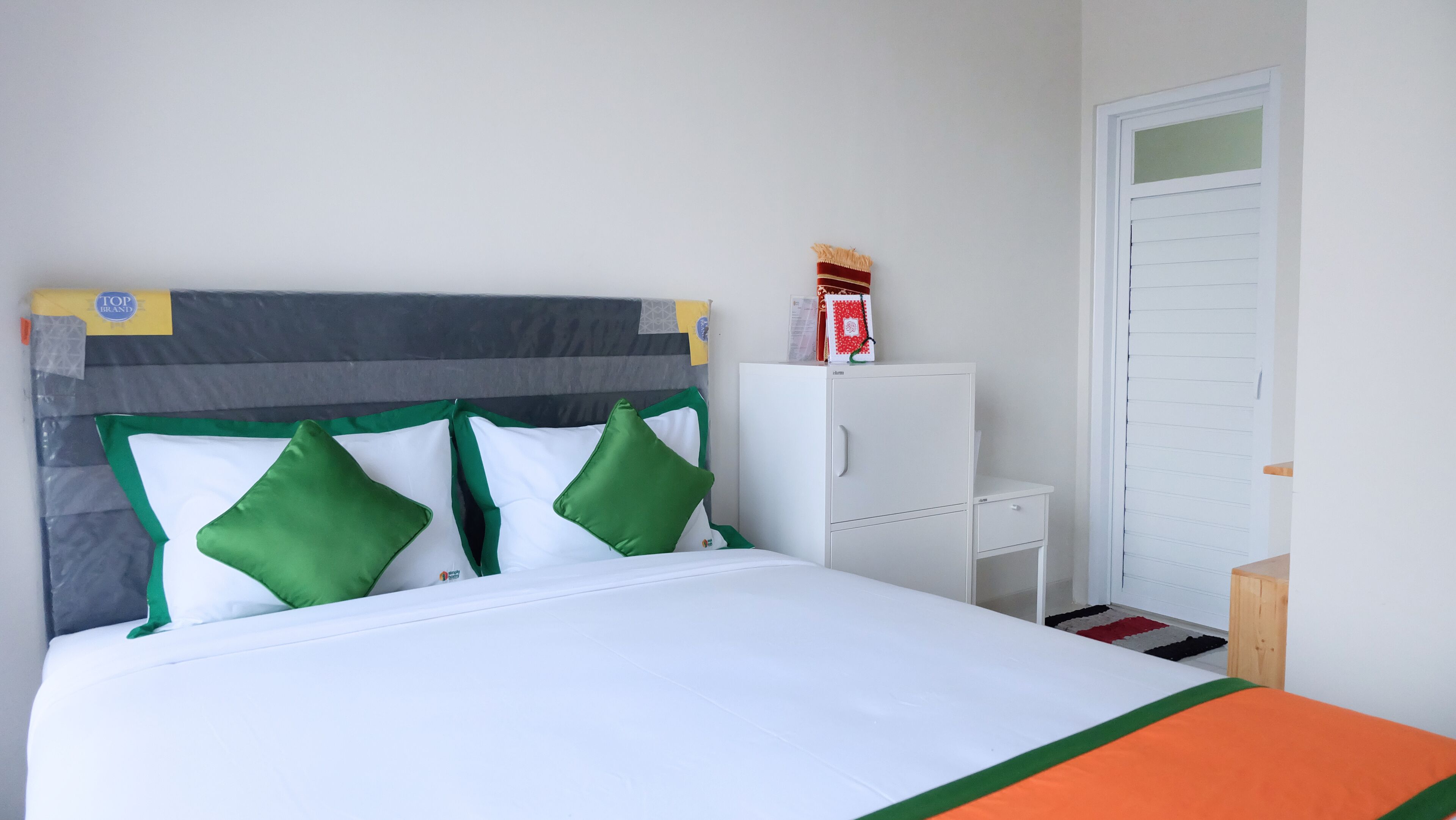 Een netjes opgemaakt bed met een grijs hoofdbord, witte en groene kussens en een witte matras. Daarbij een klein wit nachtkastje met een lamp en een houten bijzettafeltje naast het bed. Tegen de muur staat een witte kast, en er is een witte deur in de achtergrond te zien.