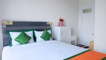 Een netjes opgemaakt bed met een grijs hoofdbord, witte en groene kussens en een witte matras. Daarbij een klein wit nachtkastje met een lamp en een houten bijzettafeltje naast het bed. Tegen de muur staat een witte kast, en er is een witte deur in de achtergrond te zien.