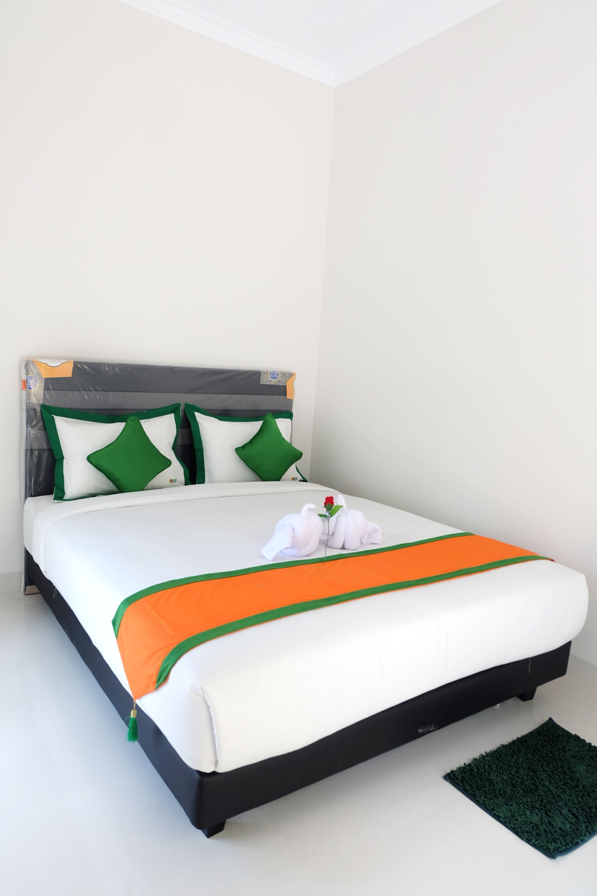 Ein ordentlich bezogenes Bett mit weißer Bettwäsche, grünen und orangefarbenen Akzenten und einem kleinen grünen Teppich auf dem Boden.