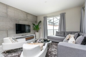 Ein modernes Wohnzimmer mit Flachbildfernseher, grauen Sofas, einem Couchtisch und einem großen Fenster mit Vorhängen.