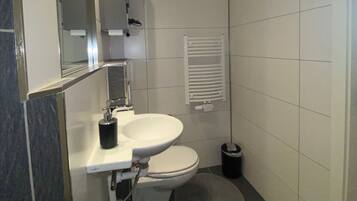 Et moderne badeværelse med en hvid vask, toilet og et spejl over vasken. Der er en håndklæderadiator på væggen og en lille skraldespand ved siden af toilettet.