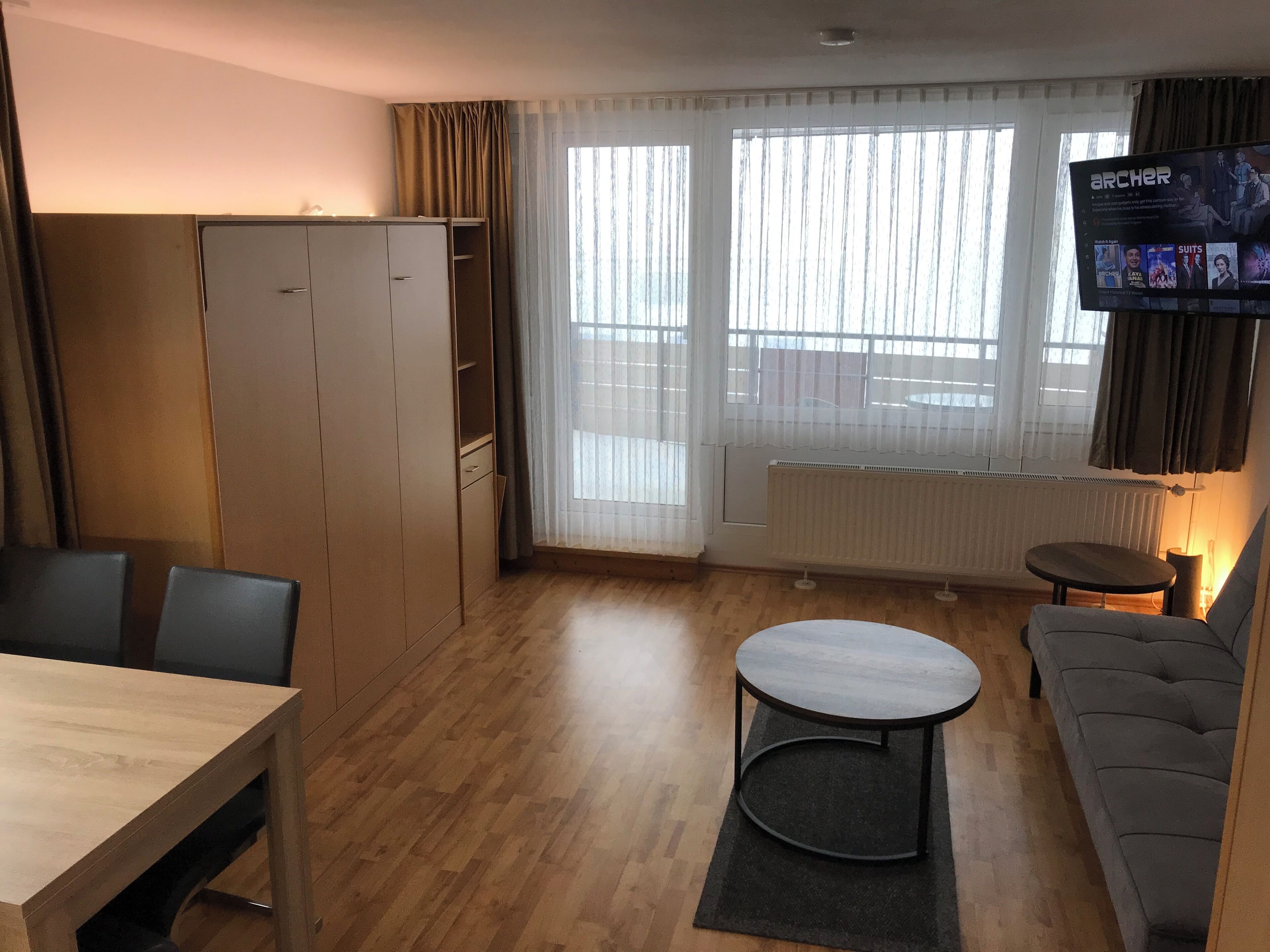 Un espacio habitable compacto con un sofá, una mesa de comedor, un televisor montado en la pared y una ventana con cortinas.