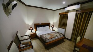 A hotel room with a bed, two bedside tables with lamps, a chair, a small table, and an air conditioning unit. - Mid Town Hotel (Islamabad)