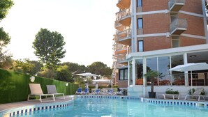 Une piscine entourée de chaises longues et de parasols, elle-même bordée d’un bâtiment en briques doté de balcons.
