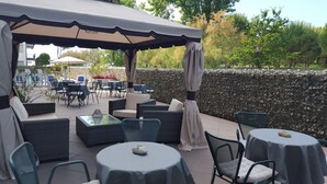 Une terrasse extérieure aménagée avec des meubles en osier, des tables rondes et un mur en pierre en arrière-plan.