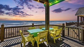 Une terrasse en bois avec des chaises jaunes et des tables vertes, d’où l’on a vue sur la plage au coucher du soleil.