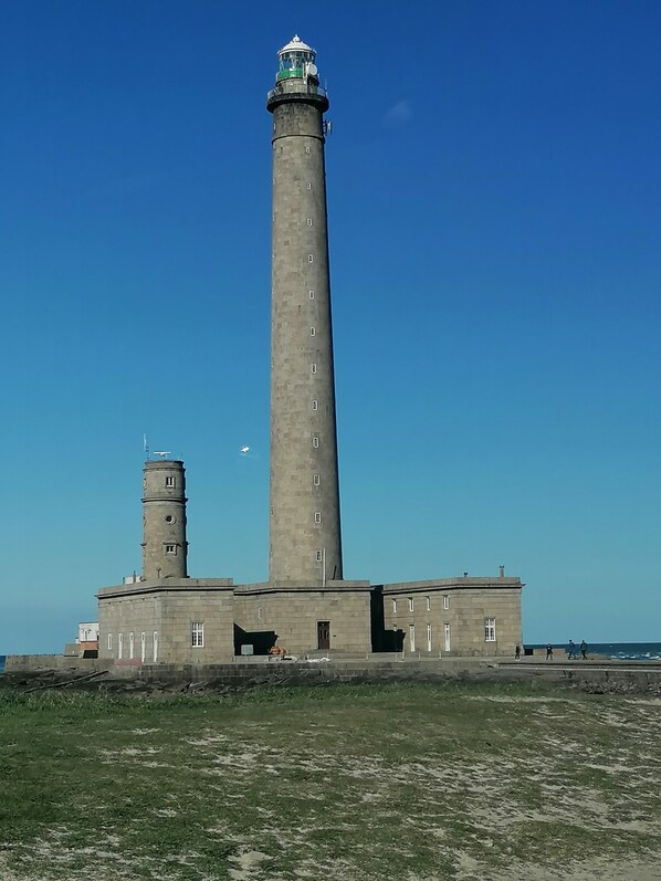 Un faro alto y cilíndrico con una torre más pequeña a la izquierda, rodeado de un área verde y un cielo azul despejado.