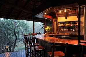 A bar area with a wooden bar counter, chairs, and a view of trees through large windows. - Waterbuck Lodge (Hoedspruit)