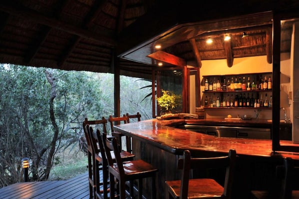 A bar area with a wooden bar counter, chairs, and a view of trees through large windows.