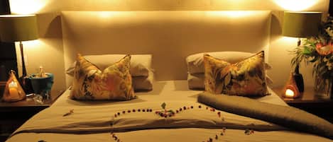 A hotel room with a bed, two pillows, a nightstand with a lamp, a vase with flowers, and a decorative item on the bed.