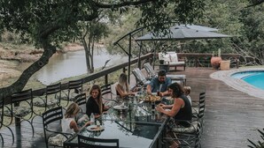 Un grupo de personas cenando en una mesa de vidrio sobre una terraza de madera, con sillas, y vistas a un cuerpo de agua.