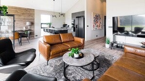 A modern living room with a leather sofa, a coffee table, and a TV. There is a kitchen in the background with a dining area. - Luxury Ocean Villas (Vestmannaeyjar)