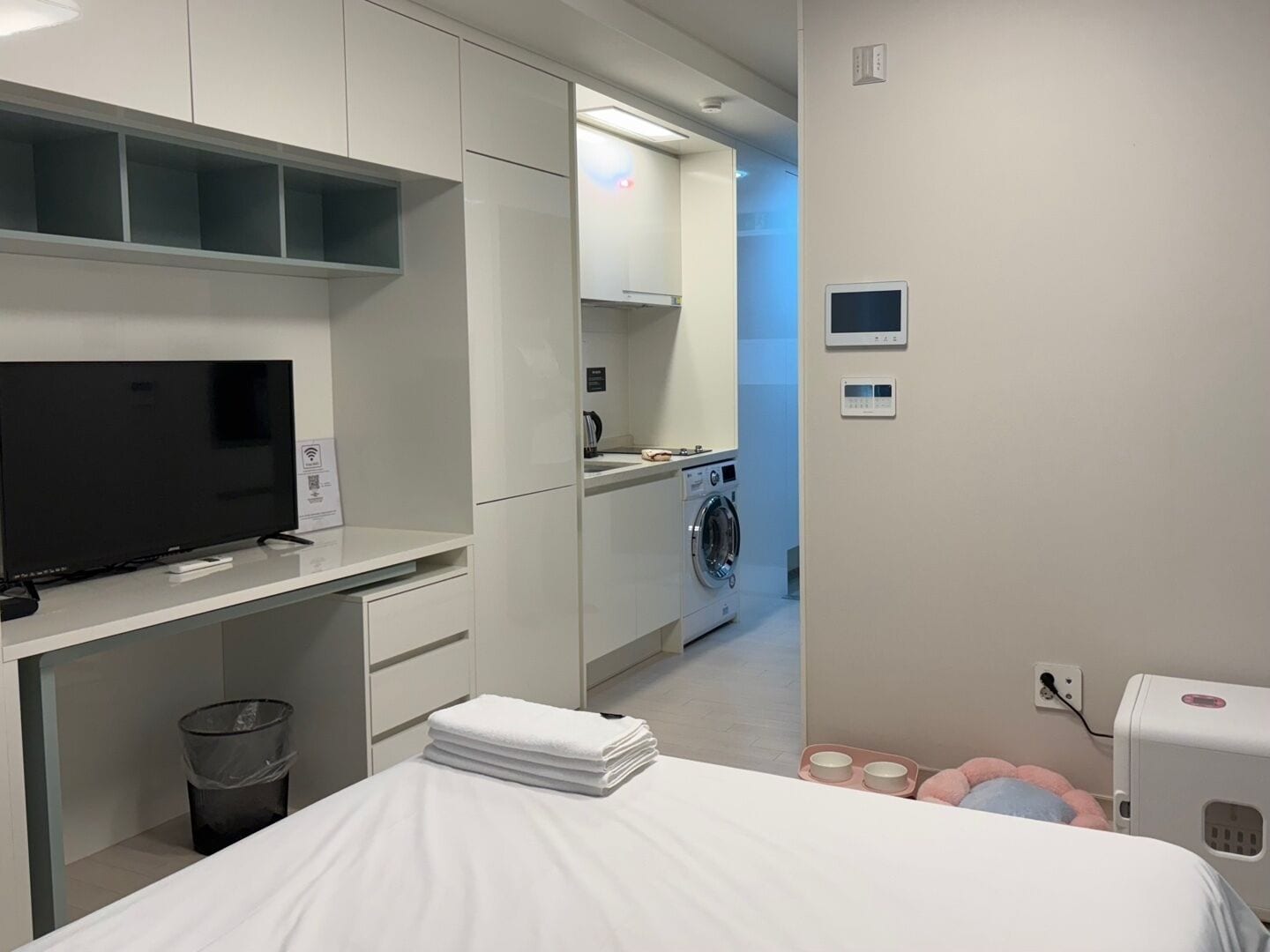 A modern hotel room with a bed, a TV, a washing machine, and a small kitchenette.