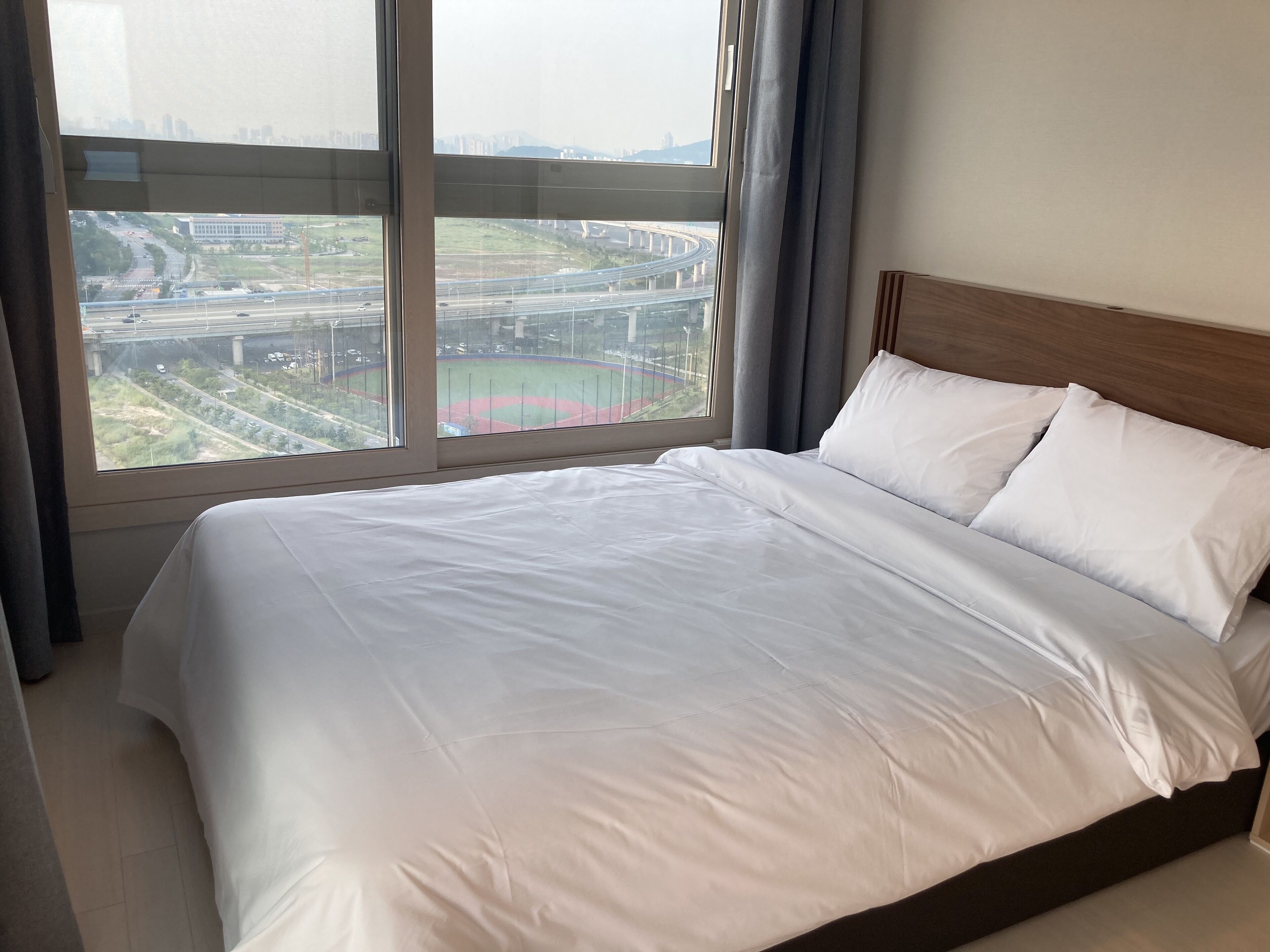 A hotel room with a large bed, a wooden headboard, and a view of a cityscape and highway through a large window.