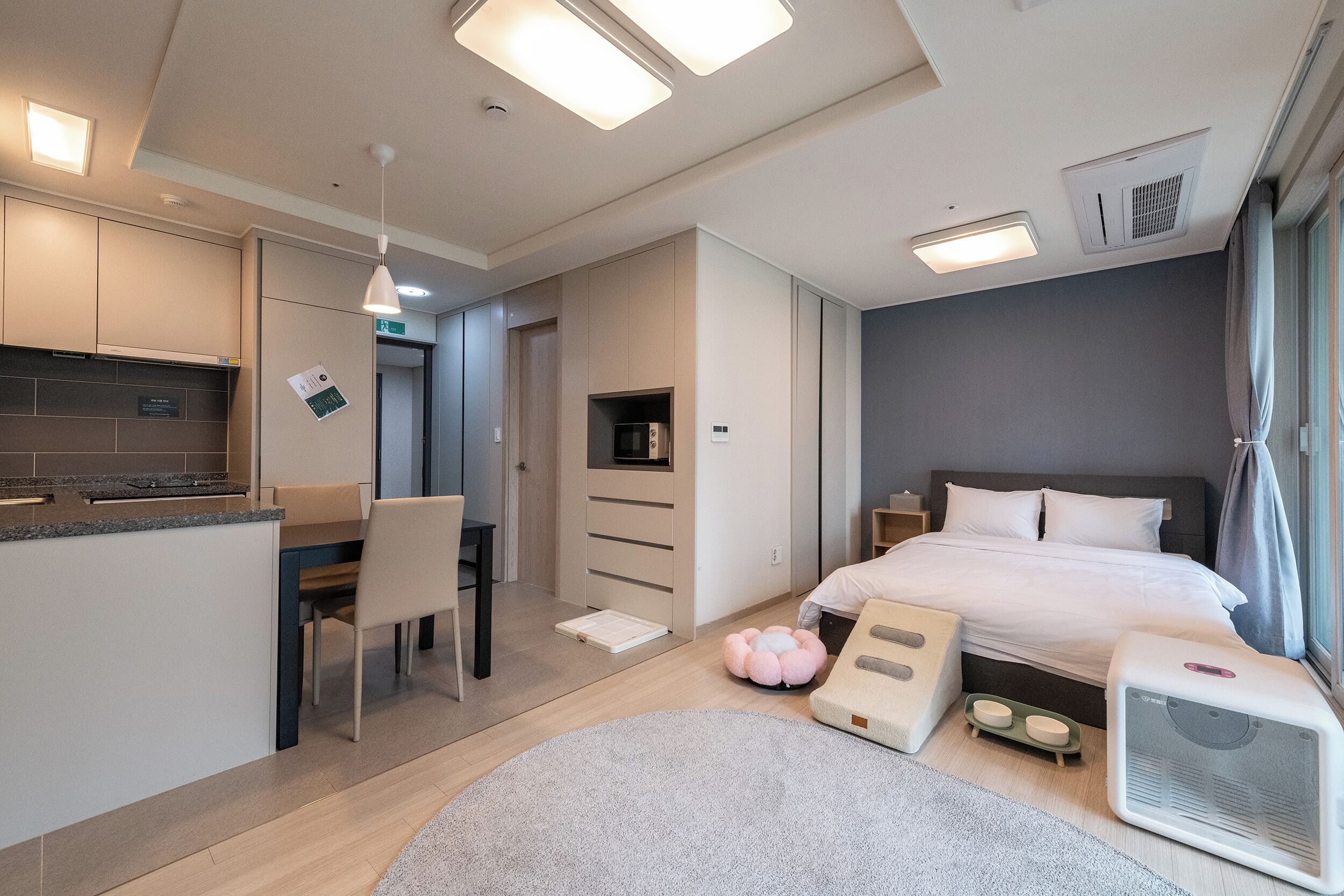 A modern hotel room with a kitchenette, a bed with a massage chair, a small table, and a bedside table with a heater.