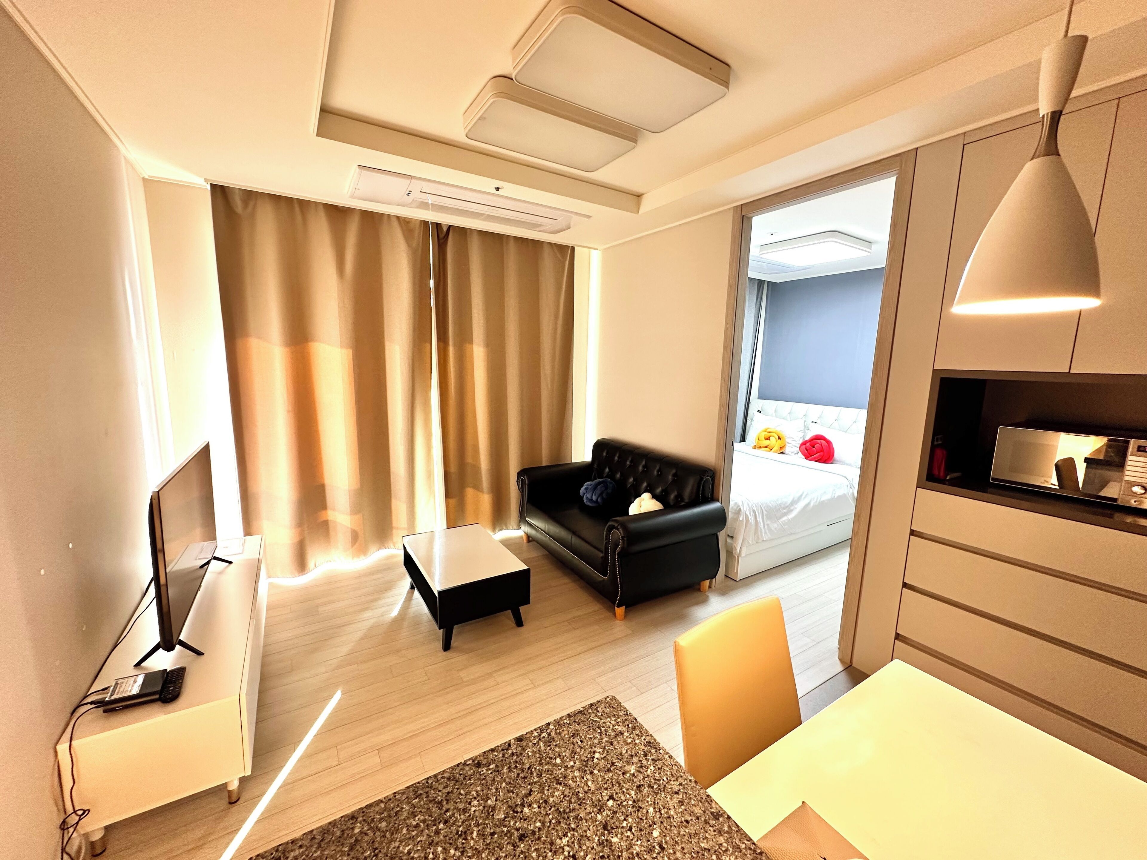 Una habitación de hotel moderna con una televisión de pantalla plana, un escritorio con una computadora portátil, un sofá de cuero negro, una mesita pequeña y una cama con cabecera blanca.