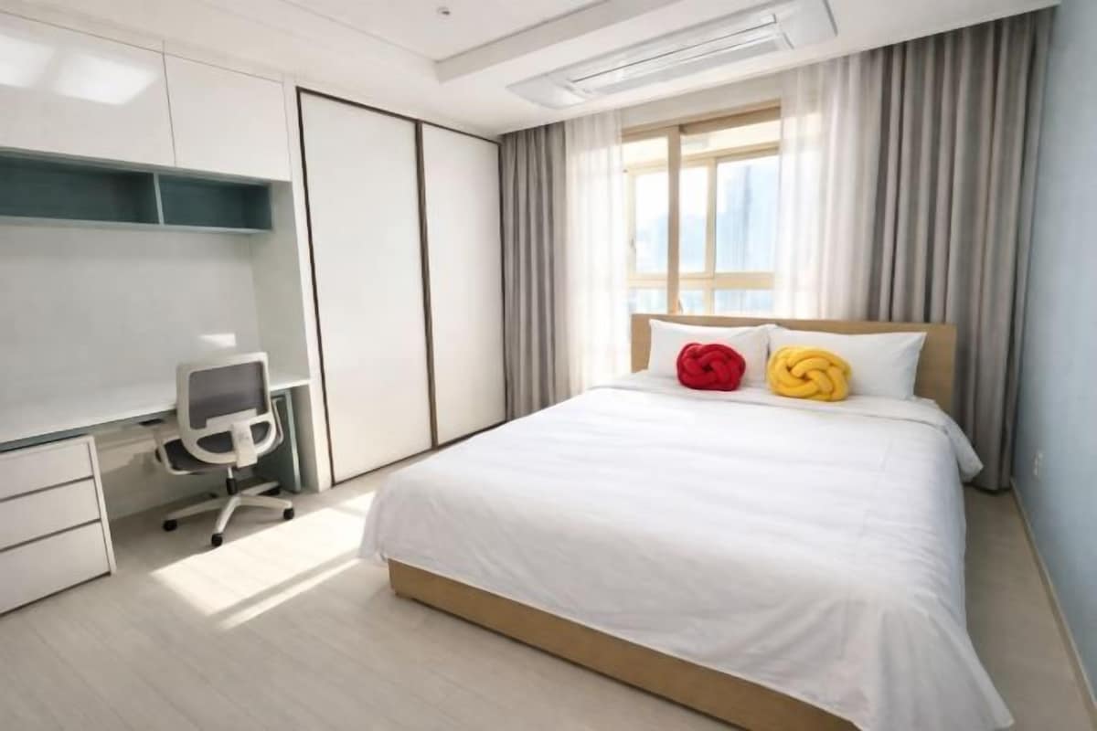 A modern hotel room with a large bed, a desk with a chair, and a window with curtains.