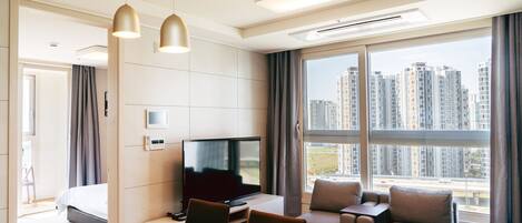 A modern hotel room with a large window, a flat-screen TV, a dining area, and a sofa.
