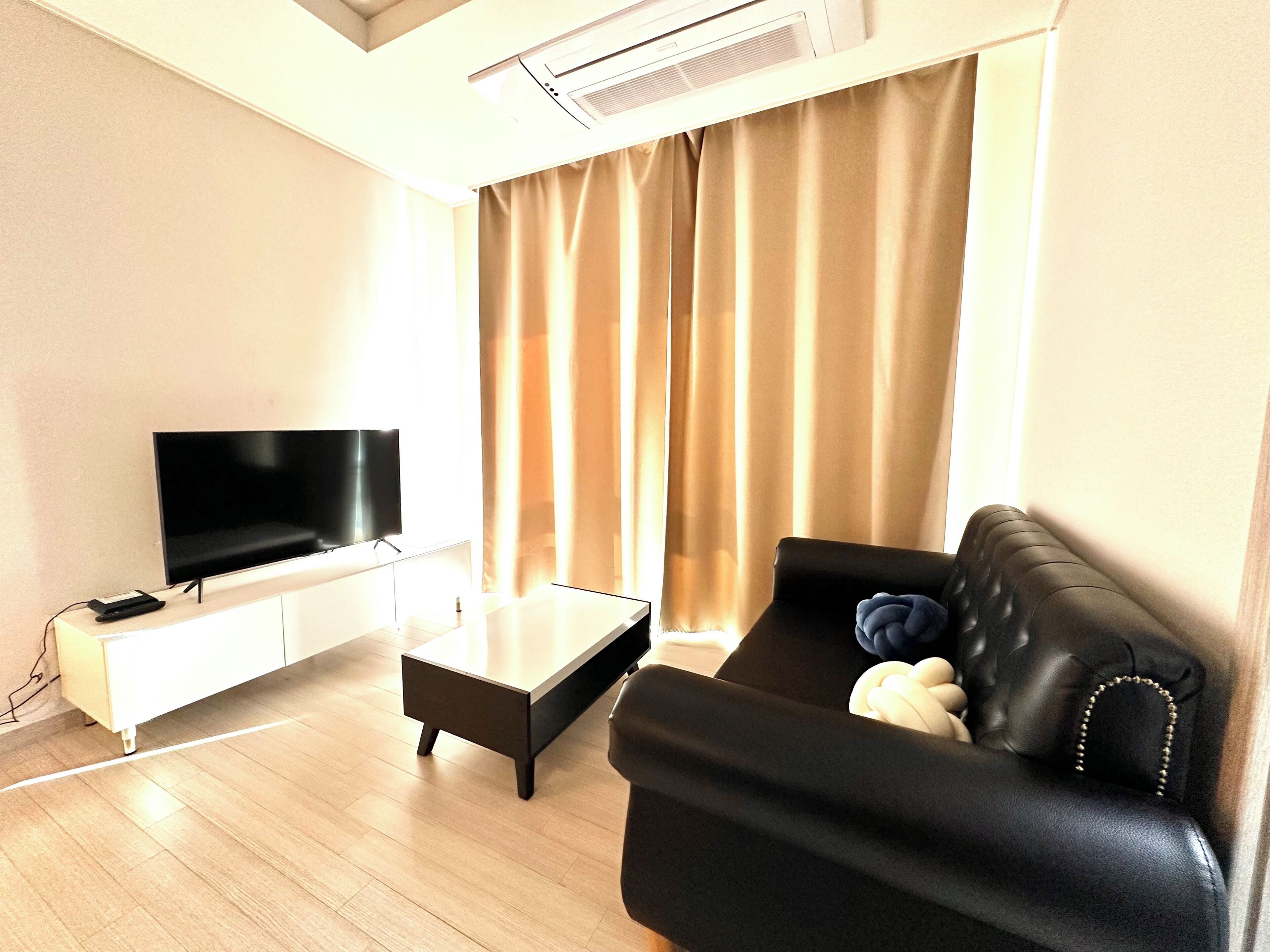 A modern living room with a flat-screen TV, a black leather sofa, a coffee table, and a large window with curtains.