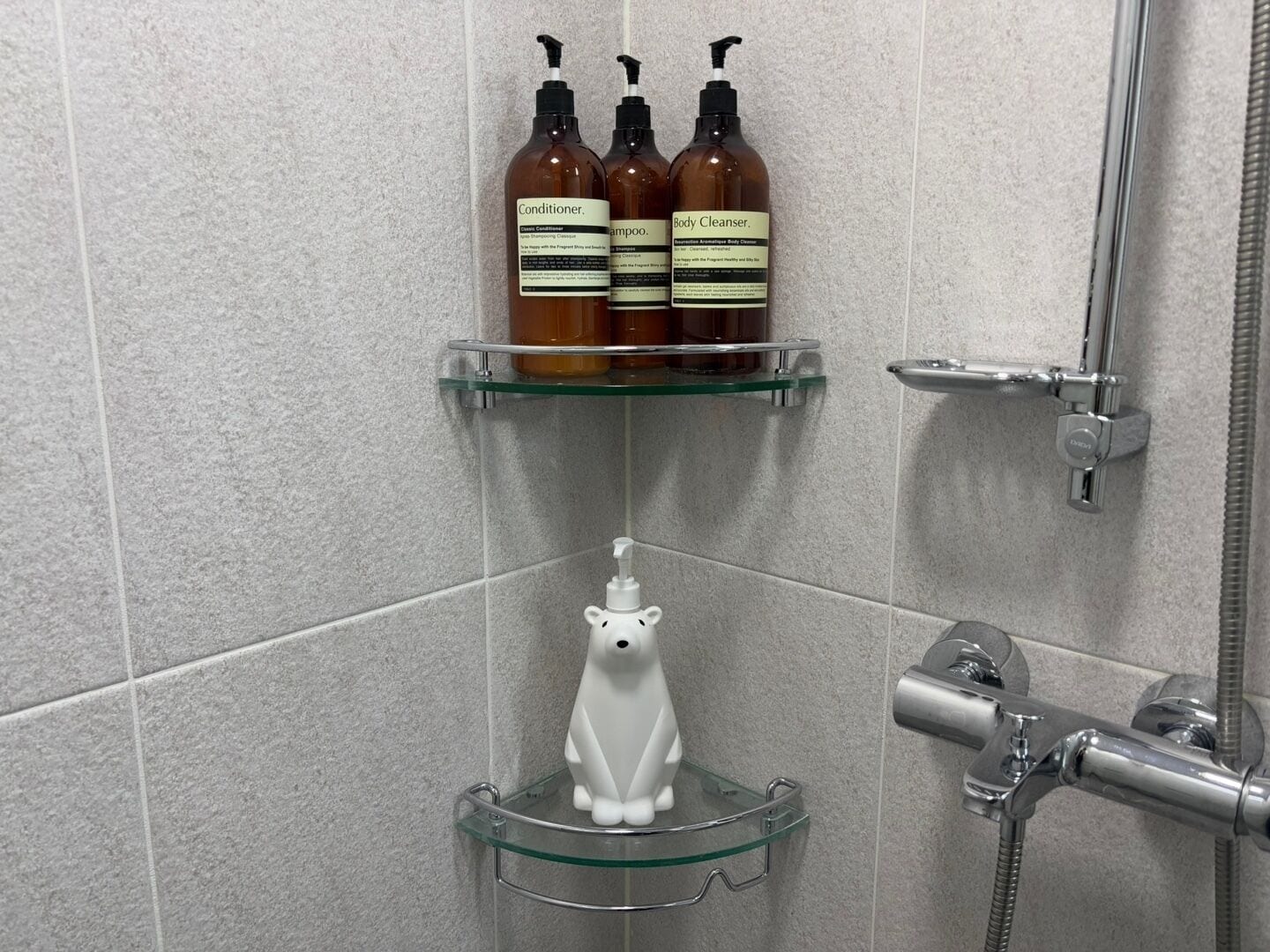 Een badkamer met drie flesjes verzorgingsproducten op een plank, een witte zeepdispenser en een douche met een handdouche.