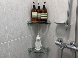 A bathroom with three bottles of body care products on a shelf, a white soap dispenser, and a shower with a handheld showerhead.