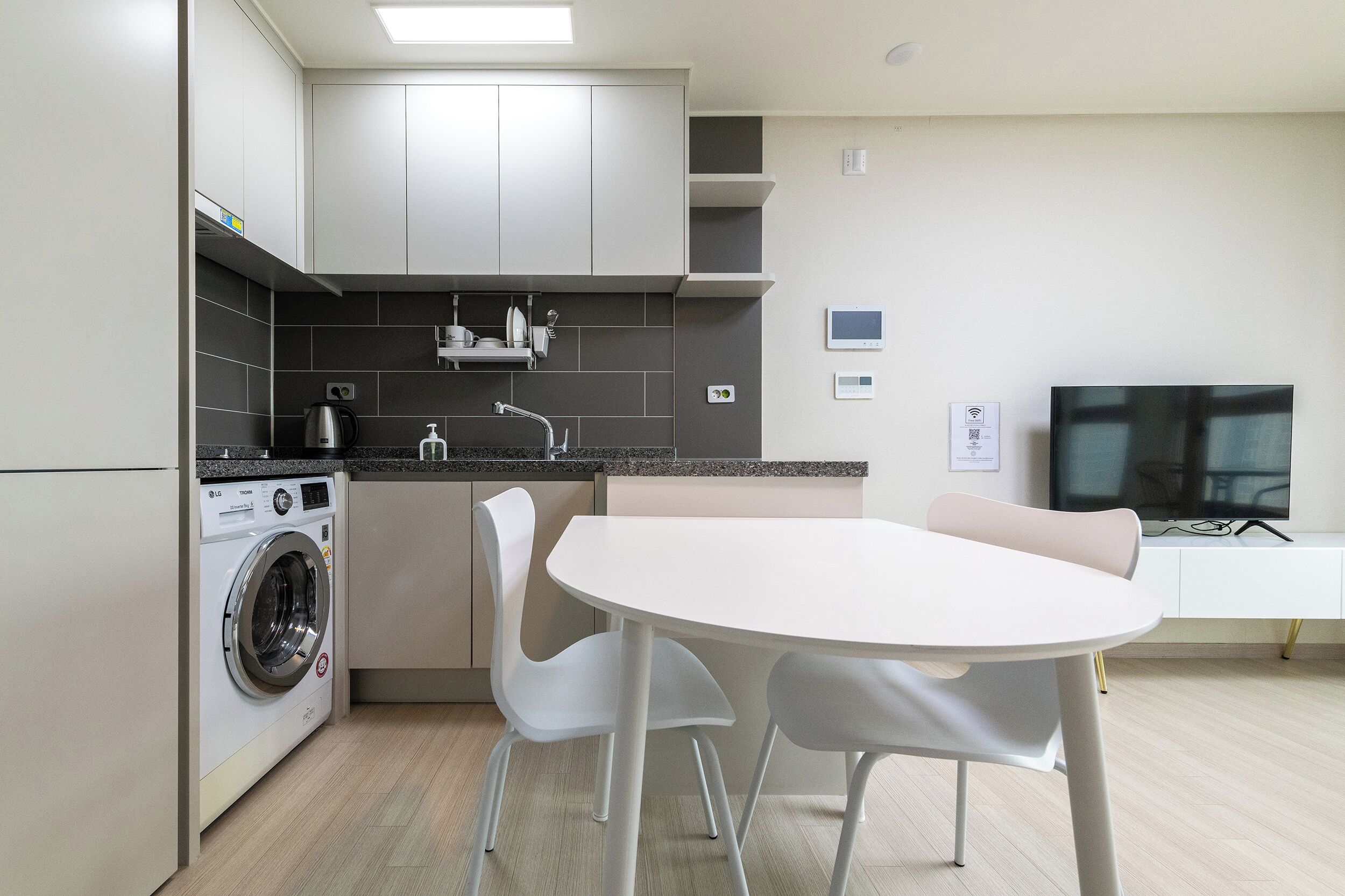 A modern kitchen with a washing machine, a dining table with chairs, and a TV on a stand.