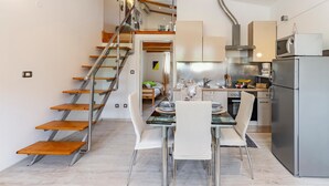 Una cocina moderna con electrodomésticos de acero inoxidable, una zona de comedor con mesa de vidrio y sillas blancas, y una escalera de madera.