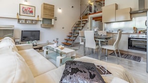 Una sala moderna con un sofá, una mesa de centro de vidrio, un televisor y una escalera que lleva a una zona de cocina.